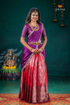 Woman in a traditional purple and red saree with jewelry, standing against a dark teal background.
