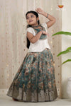 Young girl in a traditional outfit with a floral skirt and white top, posing against a light curtain background.
