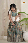 Young girl in a traditional outfit standing next to a plant with a decorative curtain in the background.