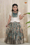 Woman wearing a traditional outfit with a floral skirt and white top, holding a leaf against a decorative curtain background.