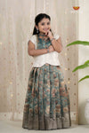 Young girl wearing a traditional outfit with a floral skirt and white top against a light curtain background.